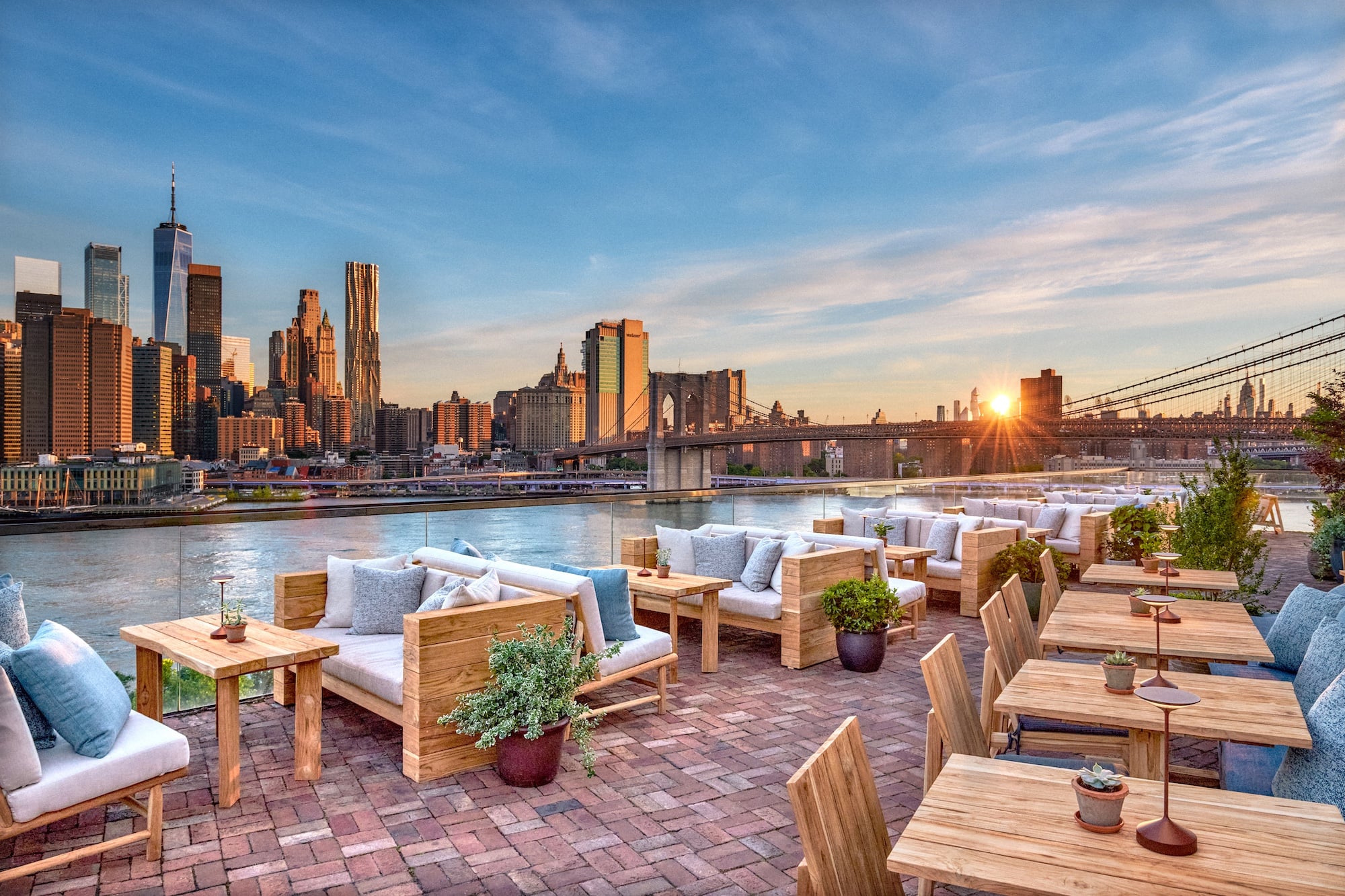 Wooden tables with comfortable cushions and wooden tables on the outside balcony with ocean and city view at 1 Hotel Brooklyn Bridge during sunset